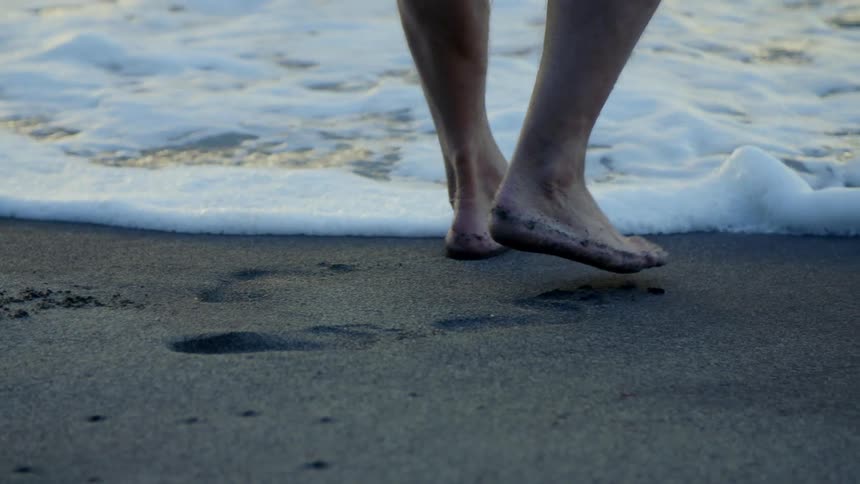 A barefoot person walking into ocean waves on a sandy beach