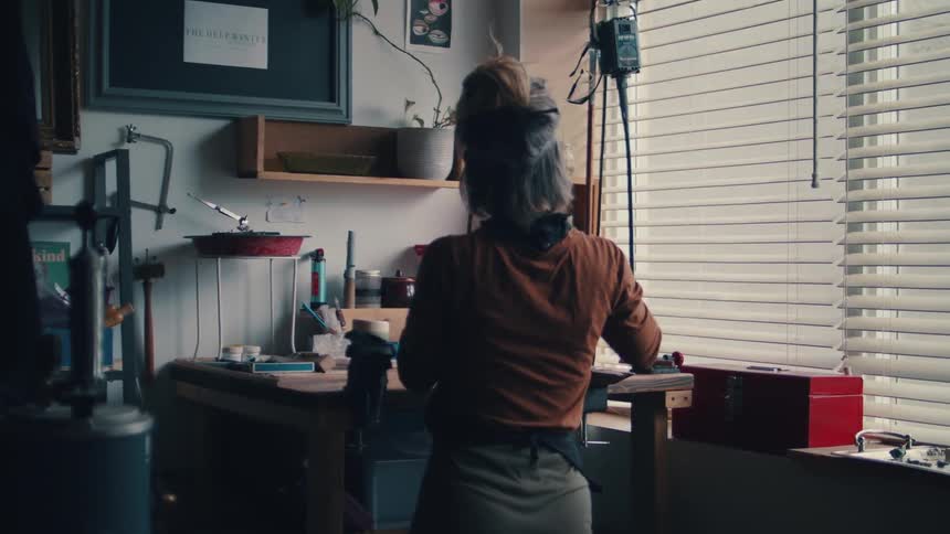 A young woman walks in to her work shop and puts her glasses on and picks up a hand saw