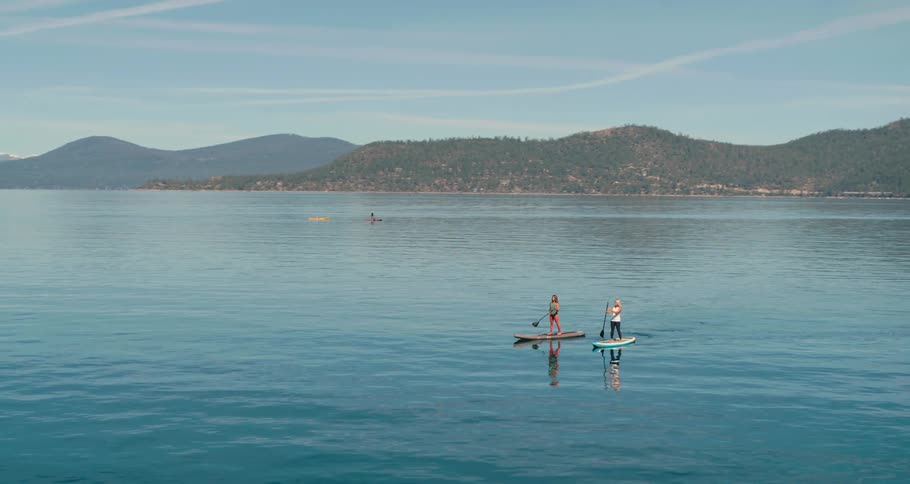 Two women paddle board across a calm lake beside a mountain