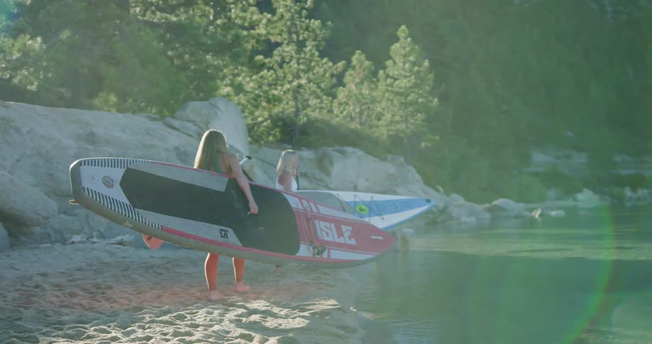 Two women carry paddle boards and oars to a lake