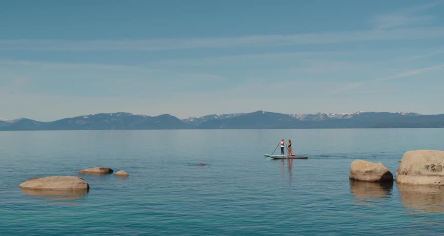 Aerial view of two women standing on paddleboards paddle through a turquoise tranquil lake against the blue sky