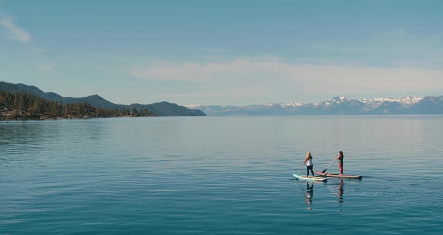 Two women paddle board across a serene lake near some mountains