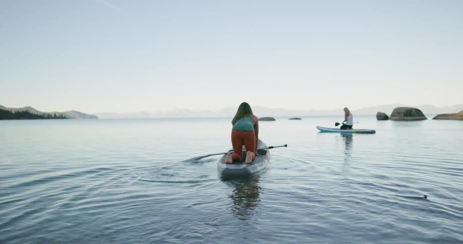 Two fit woman paddling over surfboards in a lake on the mountains at sunset