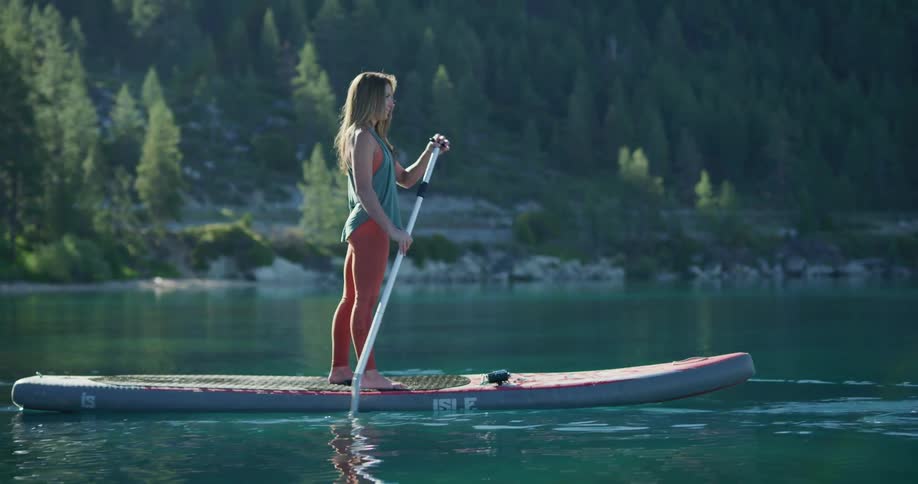 Woman paddle boards past conifer forest covered mountain slope paralleling road
