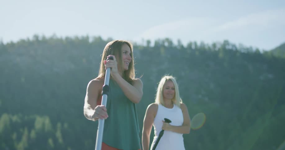 A pair of women travel side by side across a lake on their paddleboards