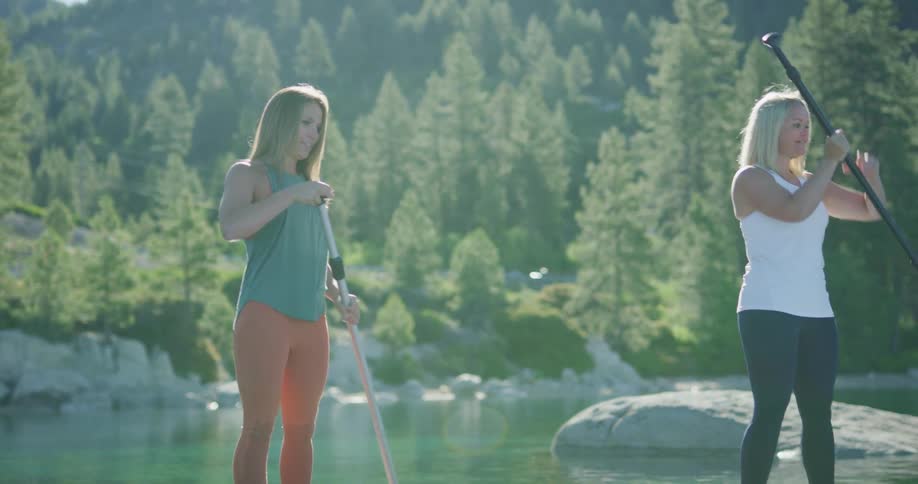 Two women uses oars to paddle board across a river