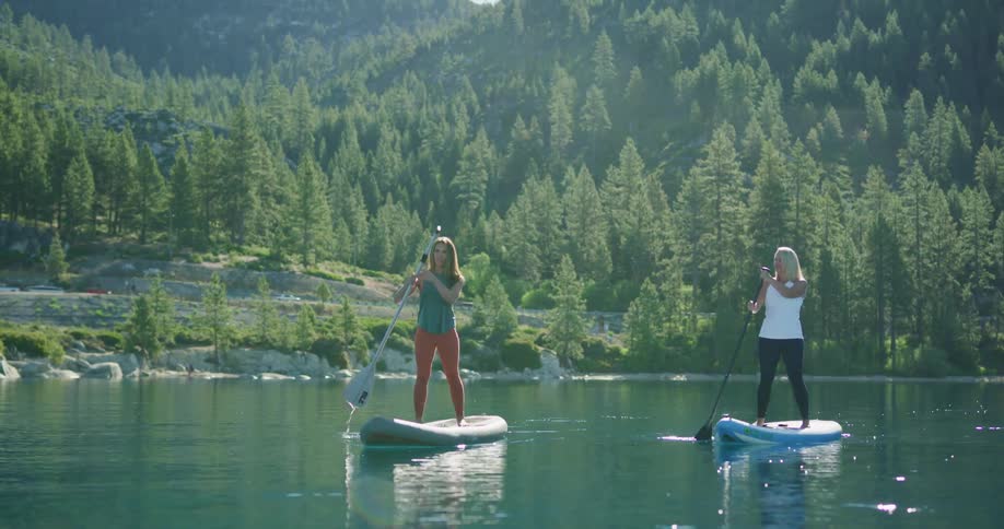 Two women are using oars to push their paddle boards on a lake surrounded by forest