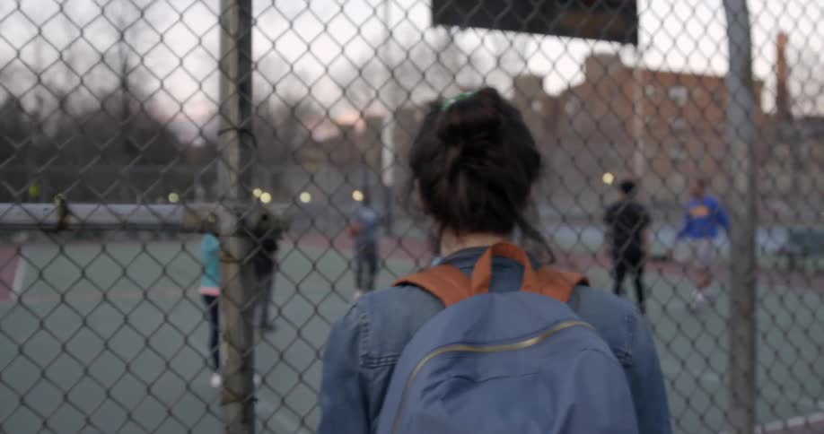 A woman drinks coffee while standing outside of a fenced basketball court