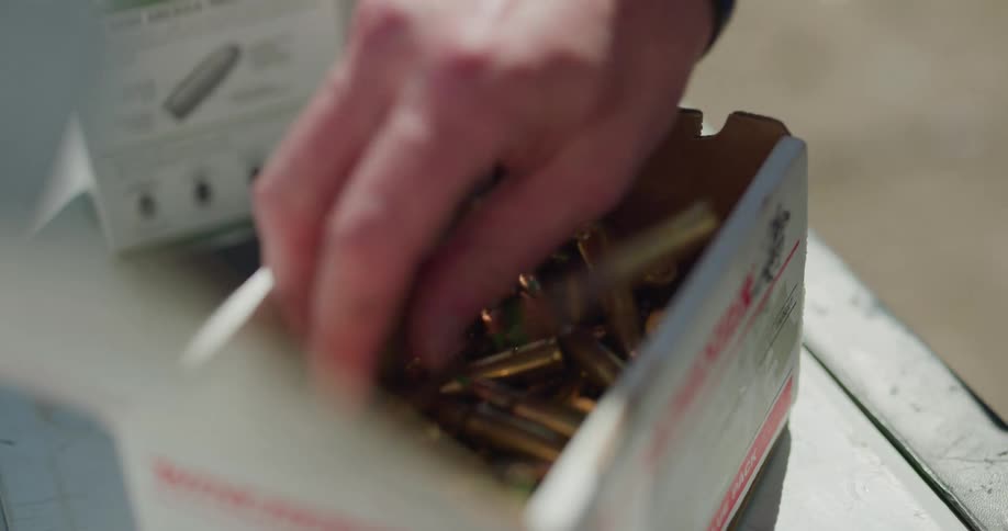 A man reaches into a cardboard box filled with rifle ammunition and picks up a few rounds