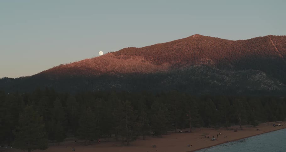 A lake with people with trees a mountain range the sky and the moon in the background