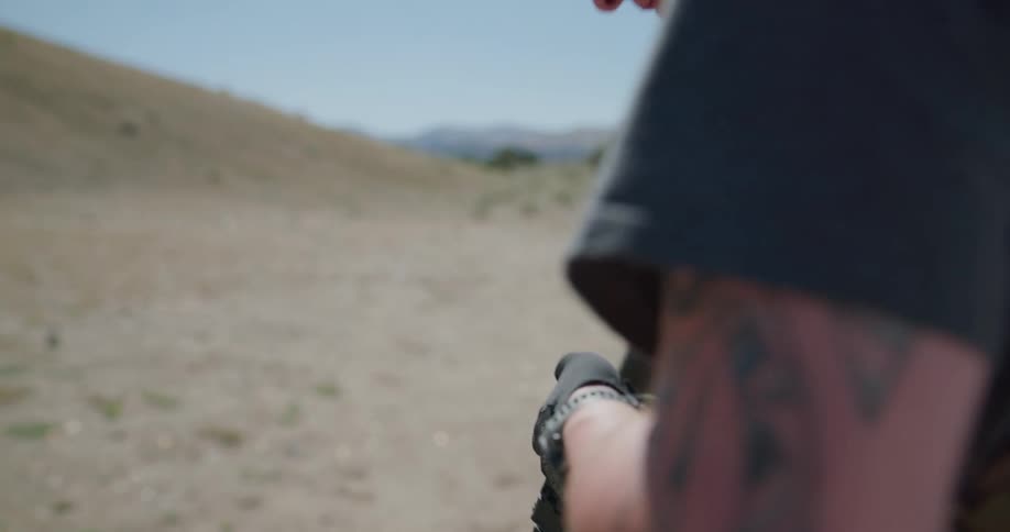 A man loads and readies a firearm while standing in a field at the base of a hill on a sunny day