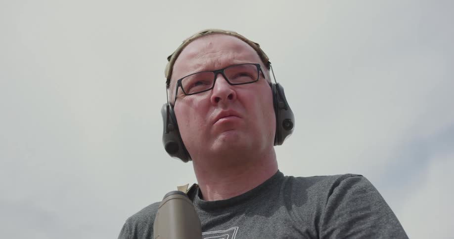 Bald man wearing glasses lowers an assault rifle and looks at the camera