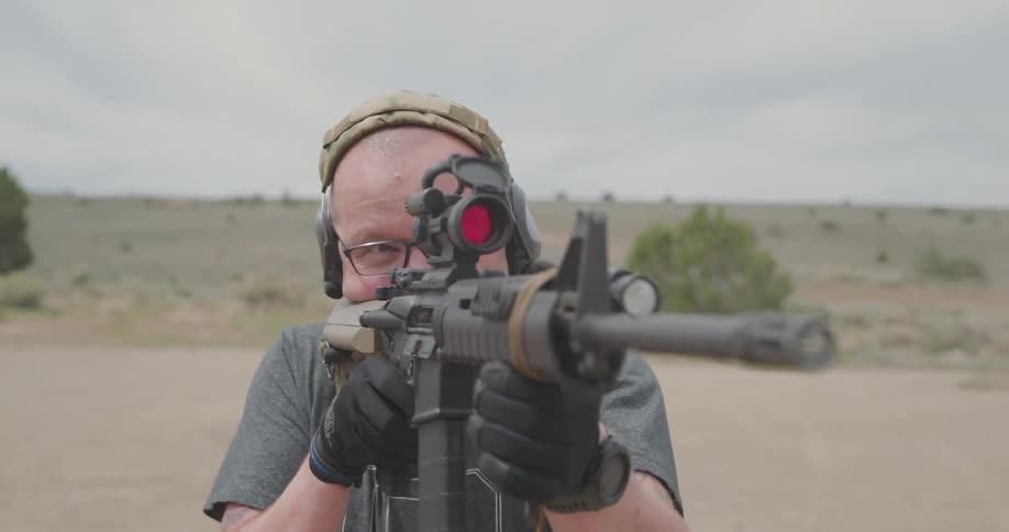 A man shoots a gun outside as a truck drives behind him in the distance