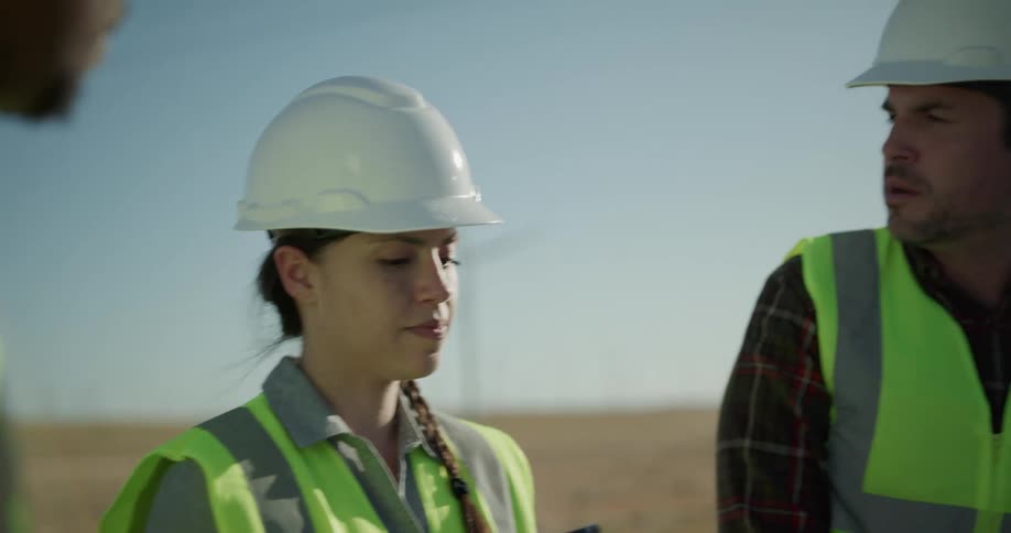 A woman holding a tablet talks to two men as they stand at a wind farm