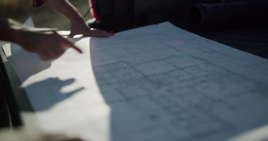 A man with a hard hat and a vest looking at blueprints in the back of a pickup truck with the sky and wind turbines in the background