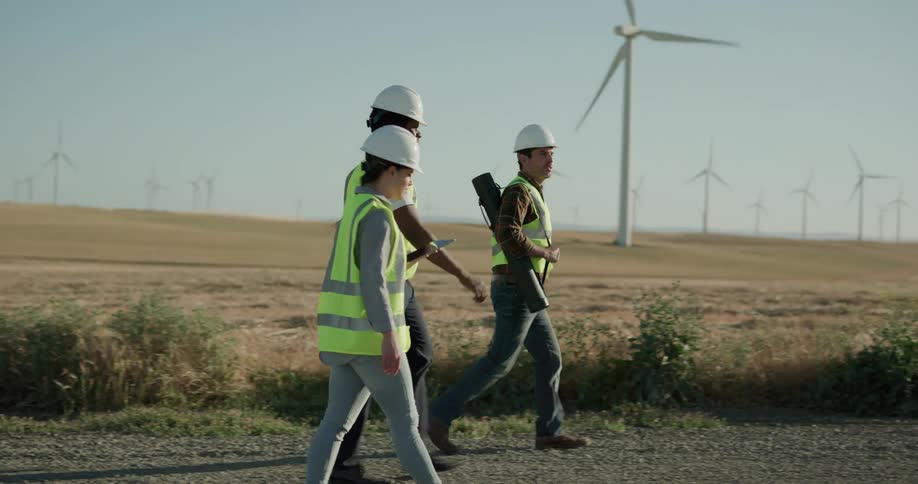 Construction workers wearing hard hats and vests walk on a gravel road next to wind turbines