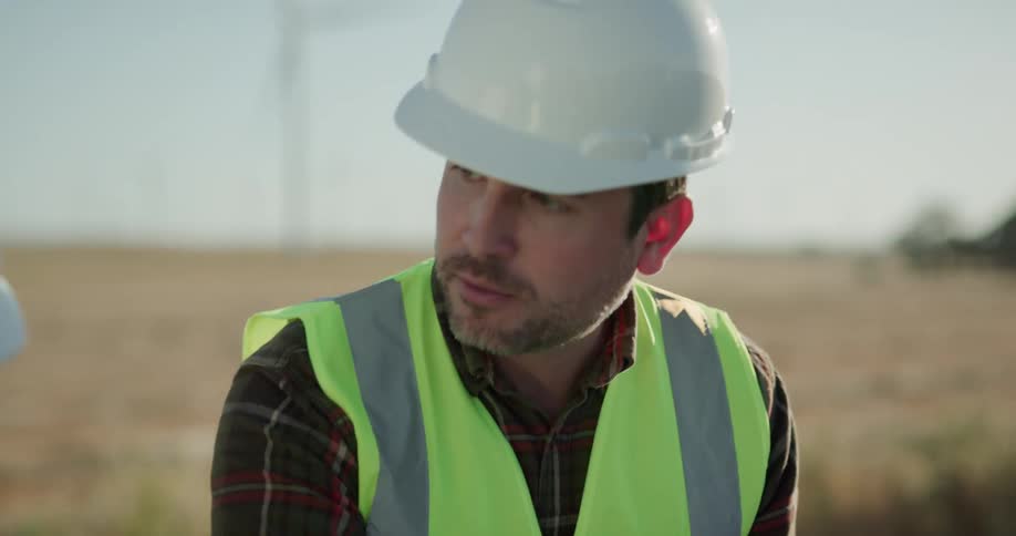 Engineer wearing hard hat and green vest talks to other workers in front of wind turbine