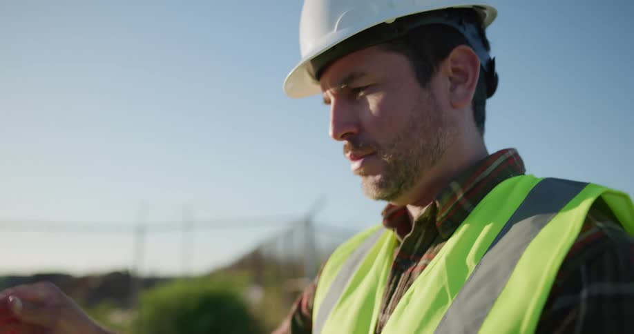 Construction worker wearing a hard hat stands outdoors while holding a technological device