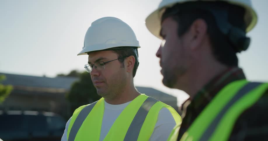 Construction supervisors wearing white hard hats and green vests talking outdoors