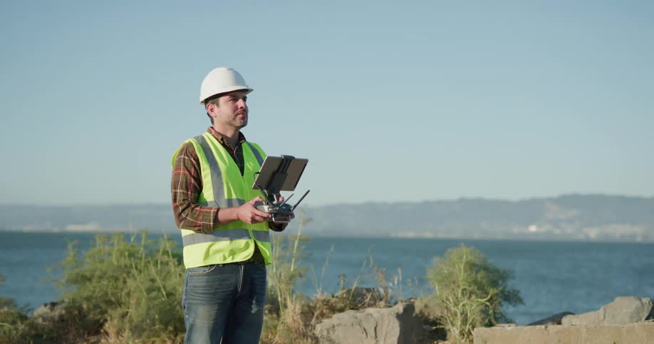 Engineer wearing white hard hat and green vest flies a drone using a remote control near the ocean