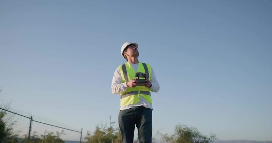 Man wearing a hard hat and safety vest holds a controller and looks up at the sky
