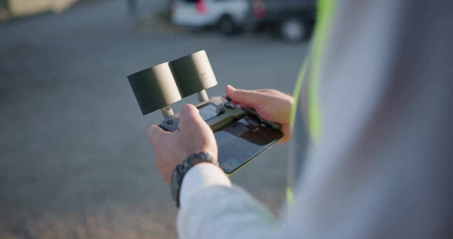 A construction worker uses a remote controller with two large antennas outdoors near a parking lot