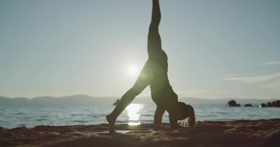 Woman does downward dog on the beach then moves into a handstand