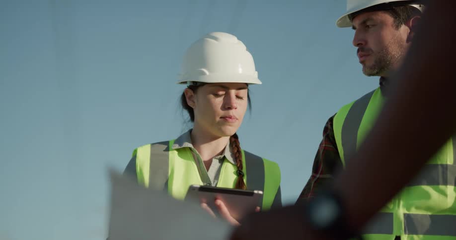 A young woman speaks and gestures at a tablet as she stands outdoors with a pair of men in similar safety gear