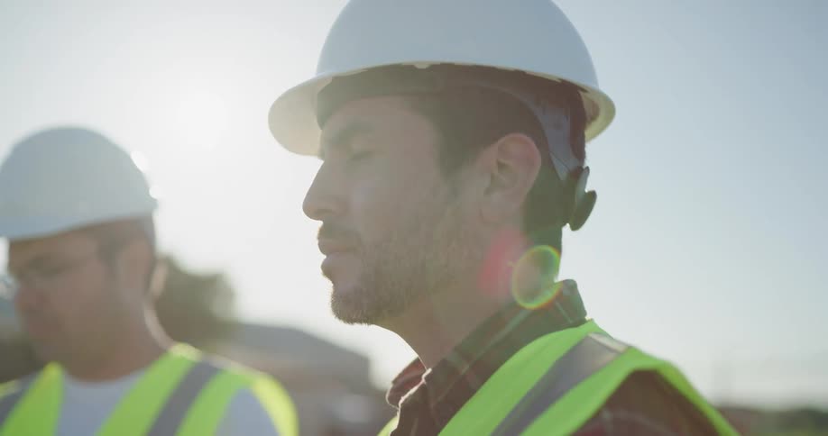 Supervisors wearing white hardhats and green vests remote controlling a drone over a construction site