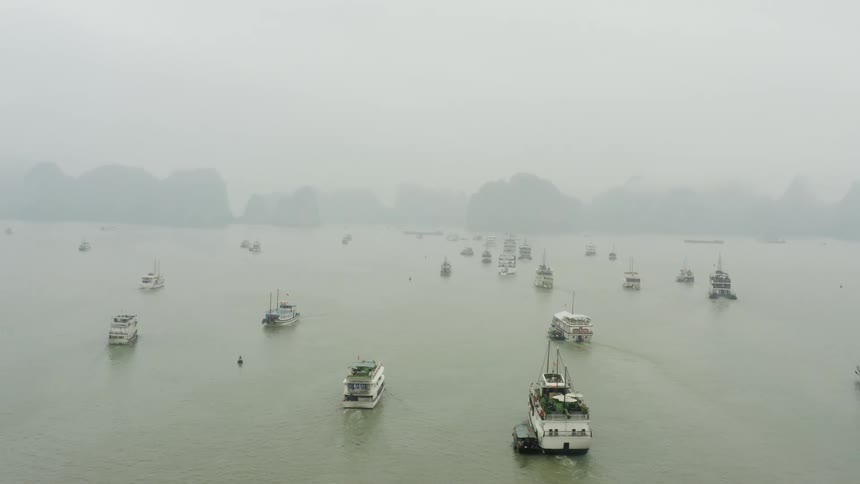 A fleet of ships and boats sail through thick fog towards a row of rock formations