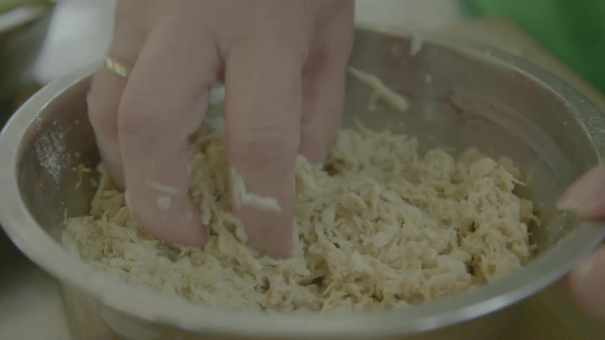 A woman mixes and stirs shredded chicken with her hand in a metal bowl