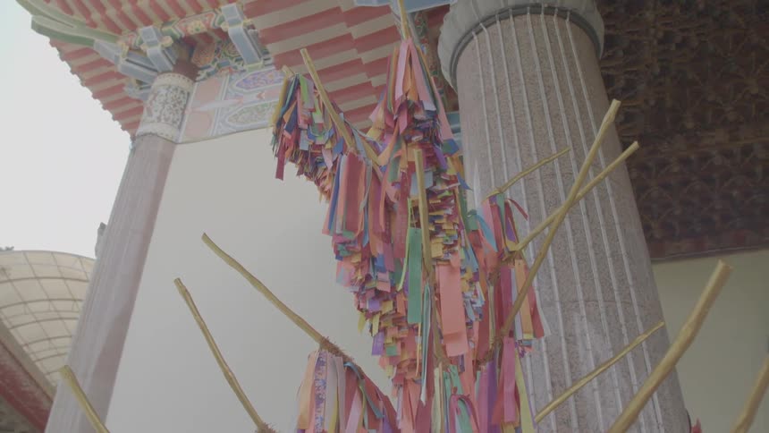 A treelike structure with golden straw branches each holding colorful hanging ribbons in front of asian architecture on a clear day