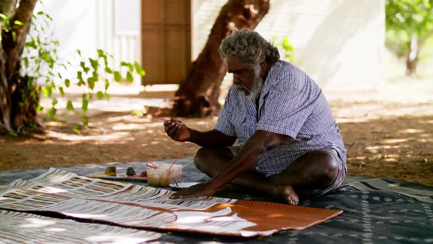 An elderly man sits on a blanket outdoors while painting on a large strip of leather