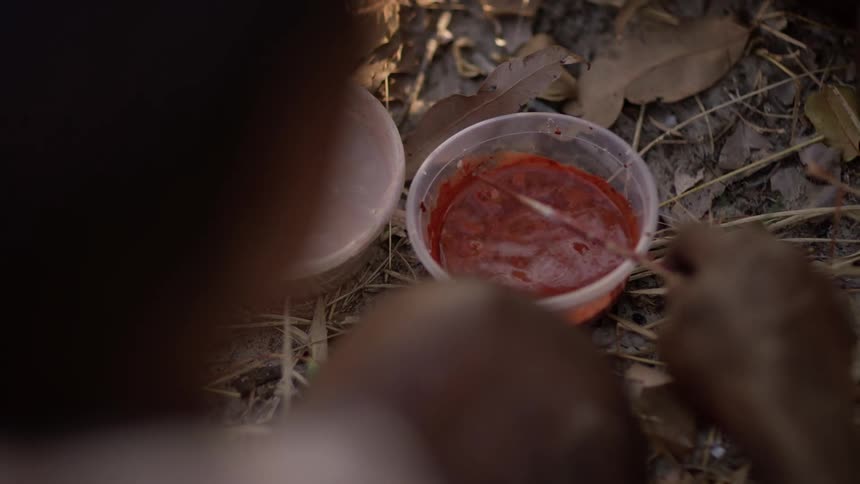 A painter kneeling in the soil dips a small paint brush into a plastic bowl filled with red paint