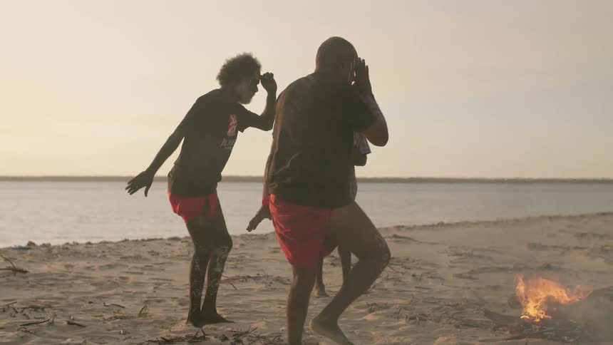 Two people in traditional loin cloth stomp and dance in the sand near a fire while another man claps