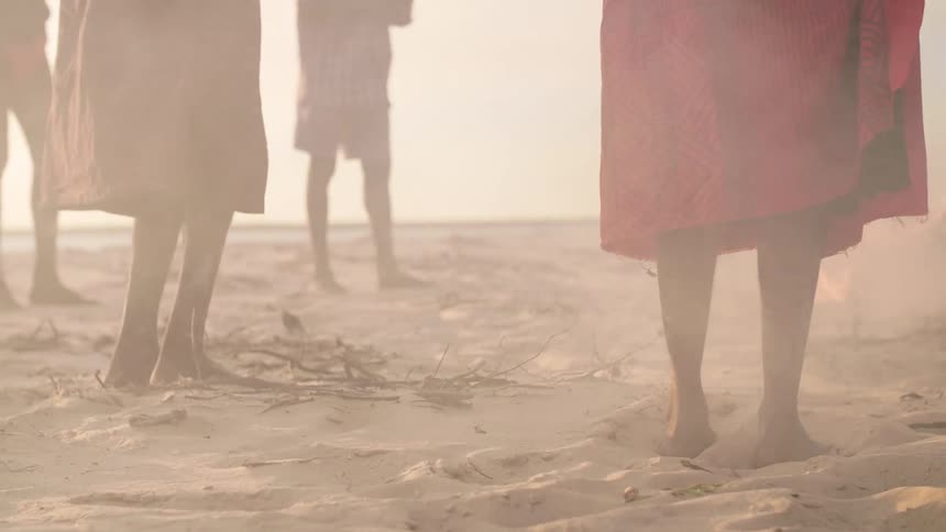 Barefoot people wearing red dance on beach near smoking fire
