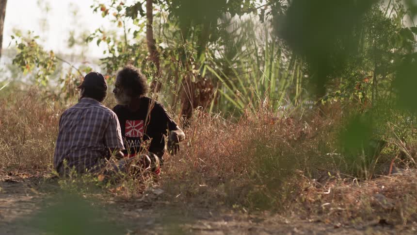 Two men sit cross legged on the ground facing each other as they talk and look at something in the distance