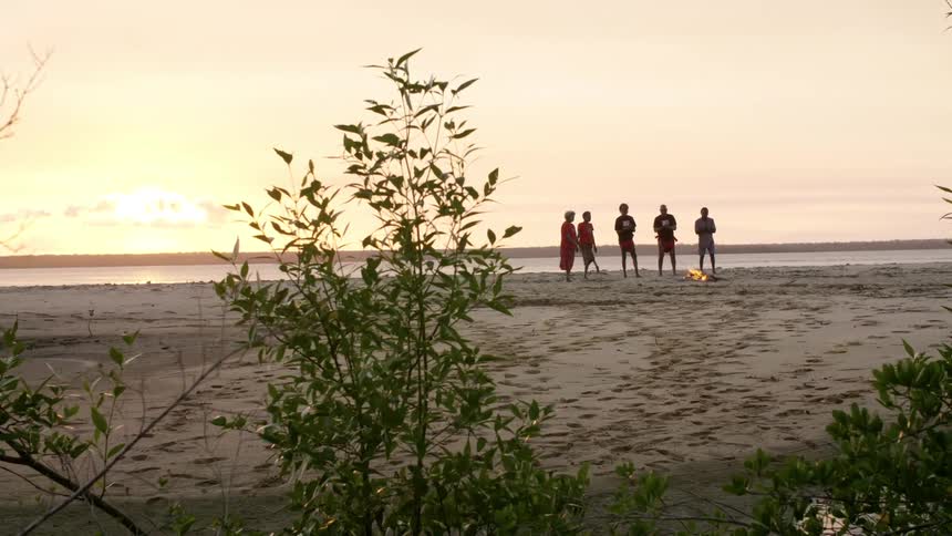A group of people clap as they stand around a campfire on the beach at dusk