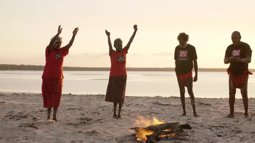 A group of indigenous people dances around a fire on the beach next to the sea at sunset