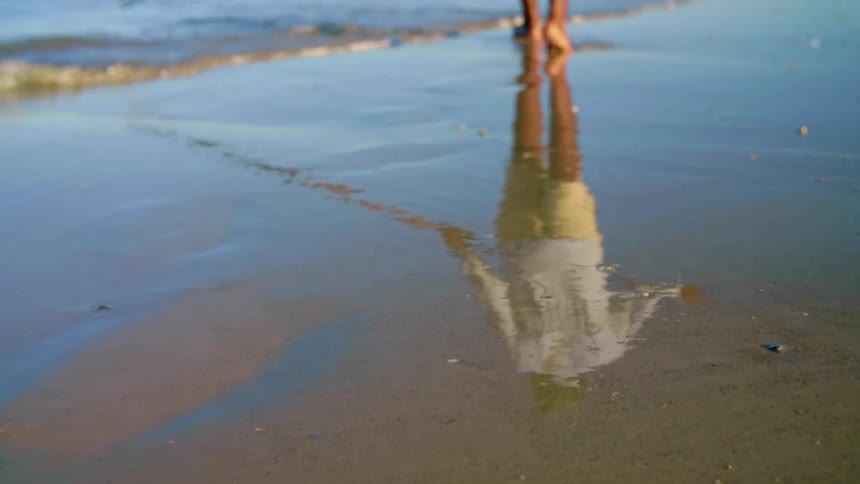 A person walks barefoot along the shallow tide on a beach during the day