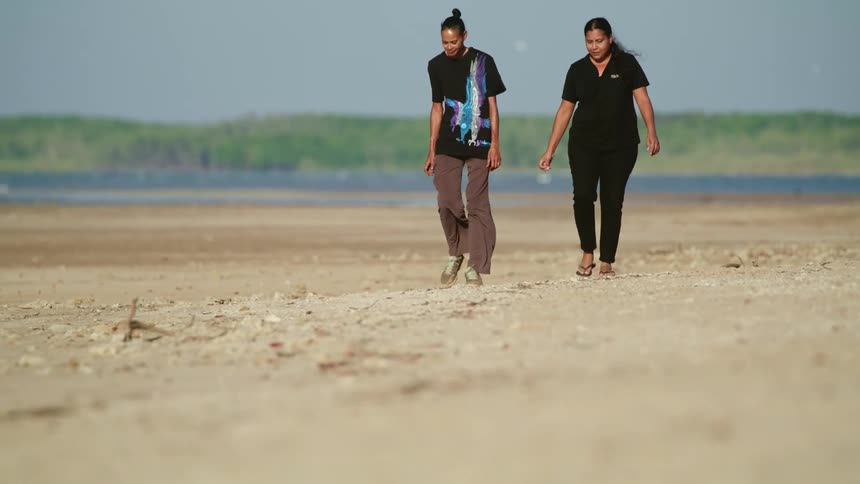 Two women walk along beach talking as one gestures
