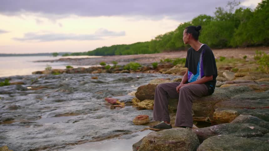 A woman sitting on a rock formation on the coast looks out over the waterfront on a cloudy evening