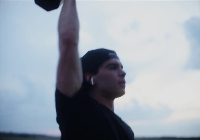 A man lifts a thick dumbbell while doing squats in a field on a cloudy evening