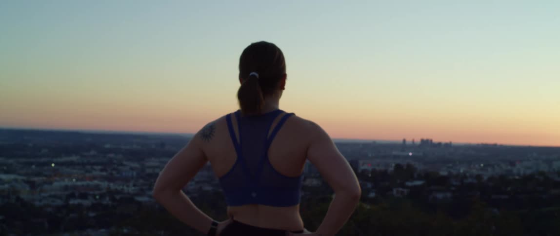Runner woman with a ponytail and sport top looks over the city from a hill at dusk