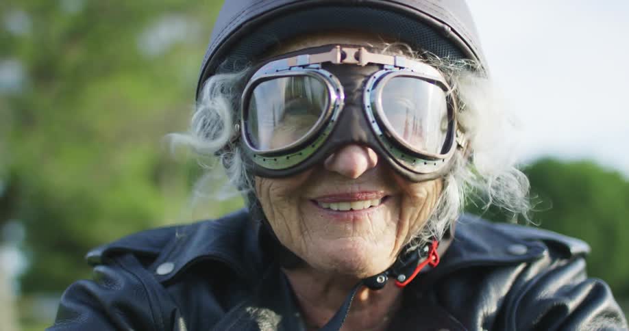 An elderly woman wearing old fashioned motorcycle equipment rides through the countryside on a sunny day