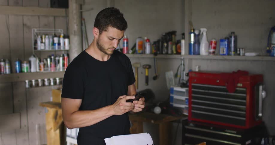 A man stands in a garage typing on a smart phone