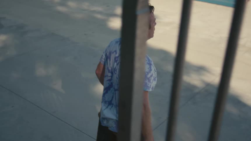 A man walking through a skate park runs his hand through his hair before dashing and jumping atop his skateboard