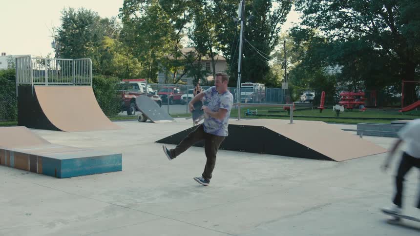 A man skateboards down a ramp in a park and bends and picks up another board and hands it to a man he skates past