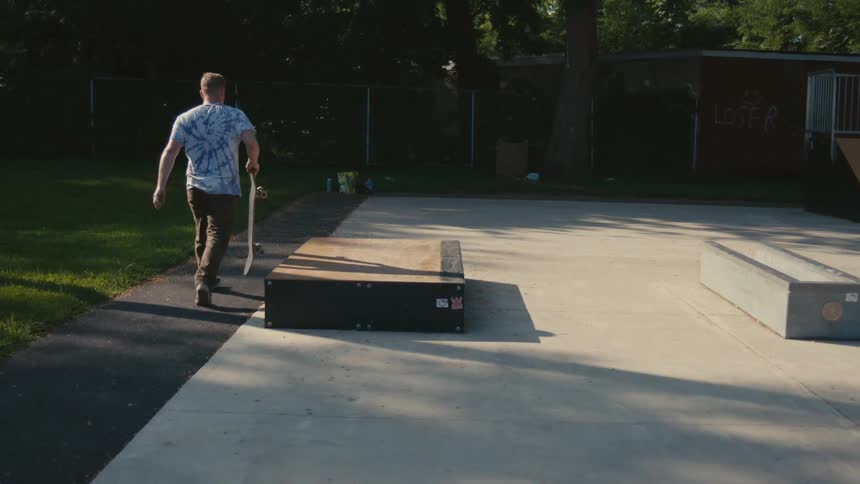 A man at a skateboard park picks up his board and walks towards a ramp where another man stands