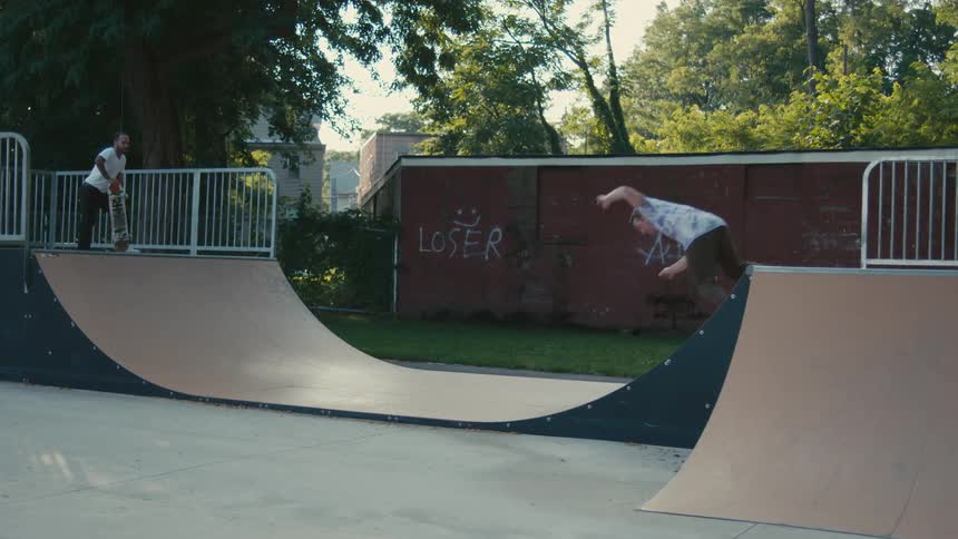 A man rides his skateboard at a skateboard park while another man waits for his turn on the ramp